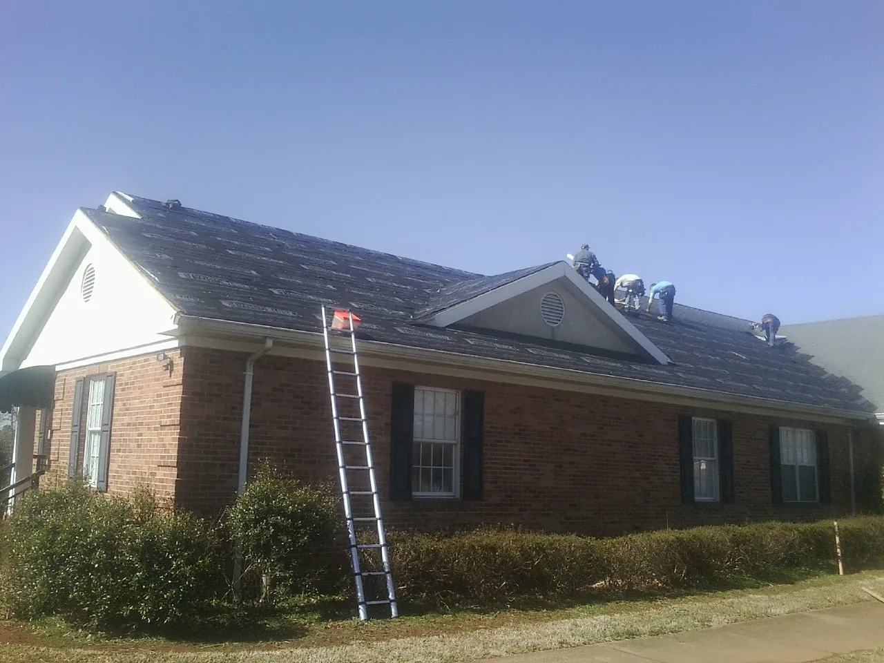 Crew tearing shingles off a brick ranch roof with a ladder set — Metal Roof Repair work by our crew in Trout Lake