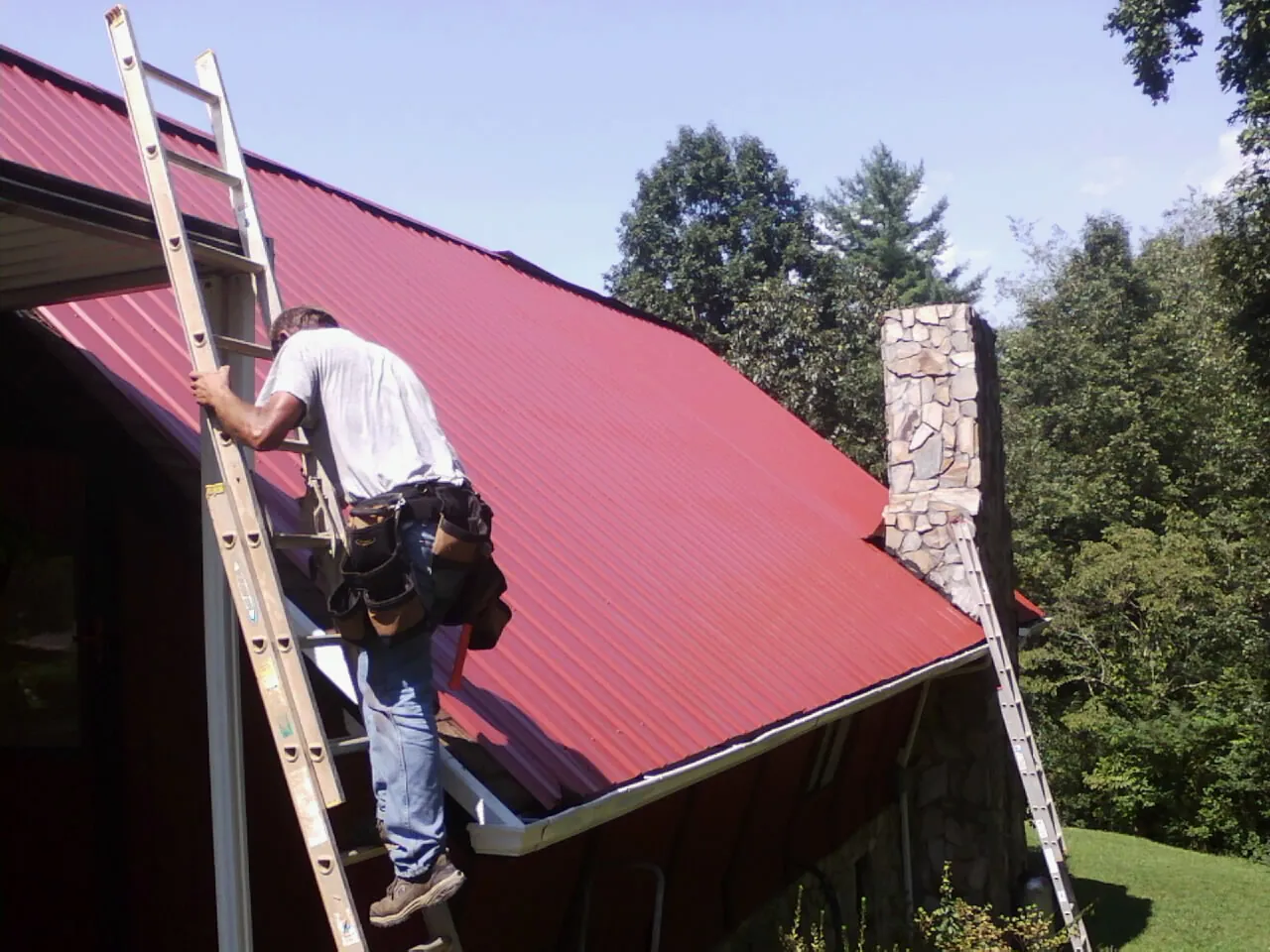 Roofer on a ladder beside a red metal roof and stone chimney — Soffit & Fascia Repair by Roofing Brothers in Trout Lake